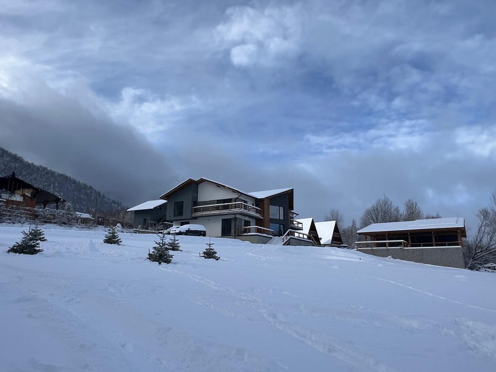 Villa Montana iarna, pe un deal acoperit de zăpadă, cu pădurea de conifere și norii peste munți